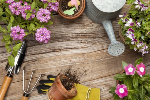 The image depicts a wooden garden table with a weathered surface, surrounded by various gardening tools and potted plants. On the table, there are two pots with flowering plants, including pink and purple blooms, alongside a metal watering can with a long spout. A small garden fork, a pair of gardening gloves in yellow, and a terracotta pot filled with soil are also visible. Additionally, a watering can with a perforated spout and a round stone container are placed on the table. The scene captures a tidy outdoor gardening space, emphasizing plant care and garden maintenance activities, typical of residential outdoor areas within Westminster or surrounding London districts. The natural lighting suggests a bright, possibly sunny day, highlighting the vibrant colours of the flowers and the textures of the wooden surface and garden tools. This setting is conducive to professional gardening or lawn maintenance services such as those offered by Lawn Mowing Westminster, supporting landscaping, planting, and outdoor upkeep in the Westminster area.
