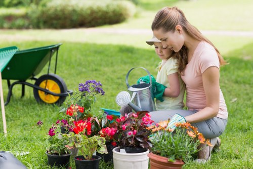 The image shows a young woman and a child gardening together in a well-maintained outdoor garden area, with a lush green lawn, flower beds filled with vibrant red, pink, and purple flowers, and a variety of potted plants. They are tending to the plants, using watering cans, with the woman gently guiding the child. In the background, there is a green wheelbarrow with a yellow wheel placed on the grass. The scene is outdoors on a bright, sunny day, with natural light illuminating the garden. The setting appears to be a residential front or back garden, landscaped with a mixture of grass, flower beds, and garden tools, supporting professional gardening and lawn care services offered by Lawn Mowing Westminster, located in Westminster or nearby areas.