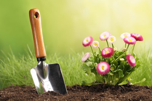 A close-up view of a garden bed showing dark, rich soil with a small silver gardening trowel featuring a wooden handle embedded into the earth. To the right of the trowel, there is a cluster of blooming flowers with pink and white petals, surrounded by green foliage. The background appears softly blurred with green hues, suggesting a well-maintained outdoor garden space likely situated in Westminster or nearby, with natural daylight illuminating the scene. The garden layout includes a freshly prepared flower bed with neat edges, positioned on a grassy lawn area, reflecting typical landscaping practices used by professional gardening services like Lawn Mowing Westminster. The scene highlights the early stages of planting or soil tending, emphasizing careful garden maintenance and landscaping efforts, which are essential components of comprehensive outdoor care in the local area.